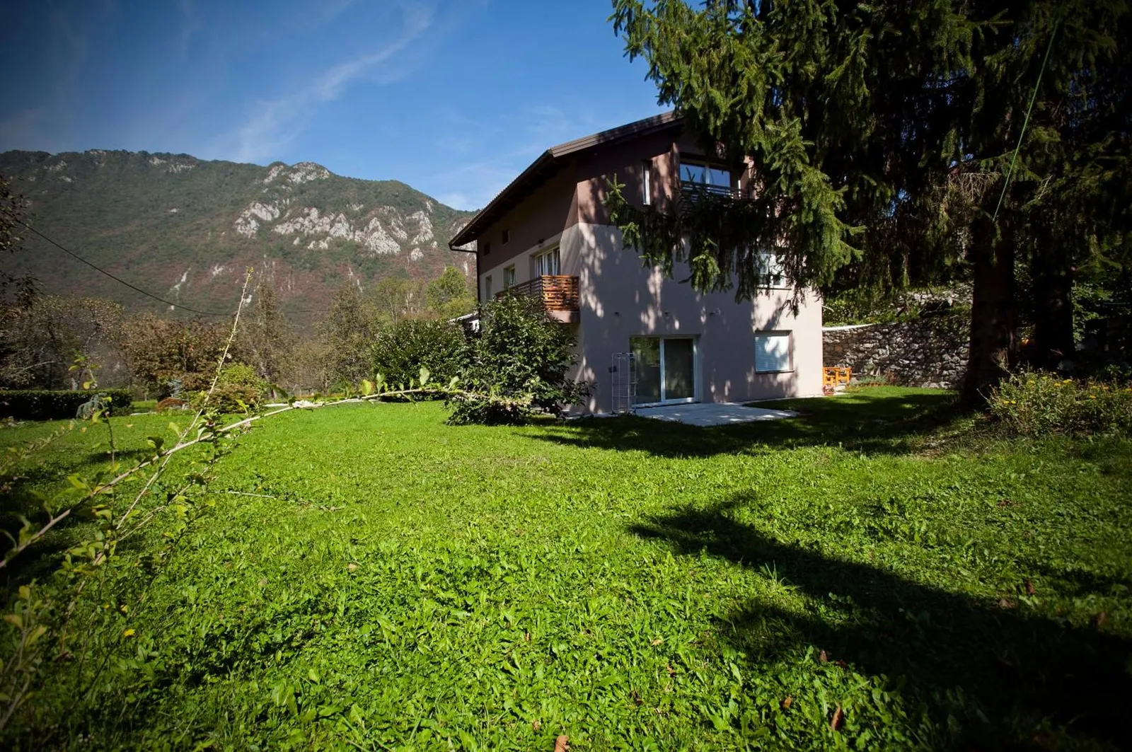 Hiša Apartmajev Soča z rjavimi balkoni, travnik pred njo in gozdnato pobočje zadaj