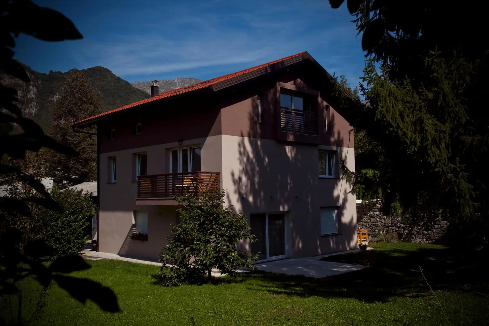 Hiša z rdečo streho in balkonom, zadaj gozdnati hribi